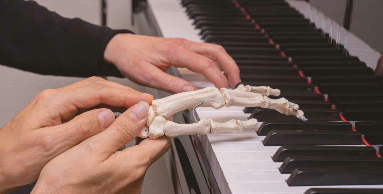 3 hender ved et piano. En hand spiller, mens de to andre holder en syntetisk skjeletthand som ligger på tangentene til høyre for den spillende handen.